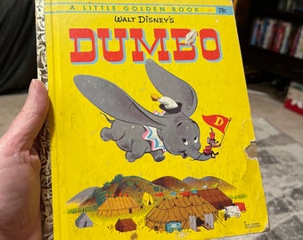 May include: A vintage Little Golden Book featuring Walt Disney's Dumbo. The cover shows Dumbo flying with his big ears, wearing a circus outfit, and a mouse holding a flag that says "D". The book is yellow with red and blue accents. The text on the cover reads "A Little Golden Book - 29c Walt Disney's Dumbo".