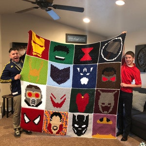Op de afbeelding: Een gehaakt deken met een raster van 20 vierkanten, elk met een ander Marvel-superheldenpersonage. Het deken wordt vastgehouden door twee mensen, één aan elke kant.