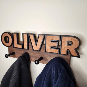 Pu&ograve; includere: Un appendiabiti in legno con il nome "OLIVER" scritto in grandi lettere con contorno nero. Le lettere sono in legno con una finitura a venatura naturale. Due ganci neri sono fissati alla parte inferiore dell'appendiabiti.