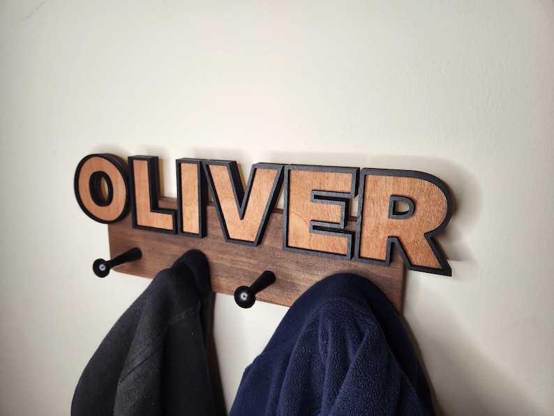 Pu&ograve; includere: Un appendiabiti in legno con il nome "OLIVER" scritto in grandi lettere con contorno nero. Le lettere sono in legno con una finitura a venatura naturale. Due ganci neri sono fissati alla parte inferiore dell'appendiabiti.