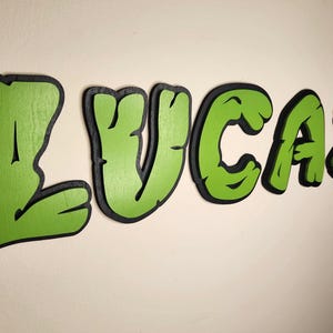 May include: Green and black wooden letters spelling out the name "LUCAS" in a cartoon style. The letters are painted green with black outlines and have a textured surface.