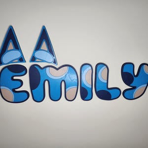 May include: Blue and beige patterned letters spelling out the name "Emily" with two blue and beige triangles above the letters.