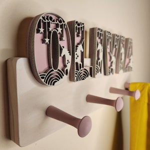 May include: Wooden coat rack with the name "OLIVIA" spelled out in pink and white letters with unicorn and star designs. The rack has three pink hooks and is mounted on a white wall.
