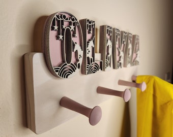 May include: Wooden coat rack with the name "OLIVIA" spelled out in pink and white letters with unicorn and star designs. The rack has three pink hooks and is mounted on a white wall.