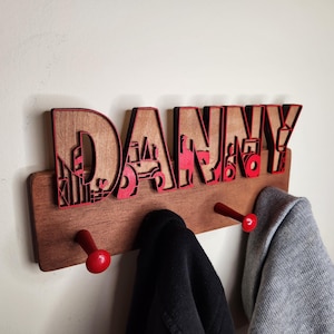 May include: Wooden coat rack with the name "DANNY" spelled out in red letters. The letters are decorated with tractor and farm-themed cutouts. The rack has two red hooks.