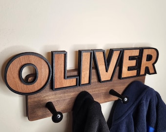 May include: Wooden coat rack with the name "OLIVER" in large, black-outlined, brown letters. The rack has three black metal hooks and is mounted on a wall.