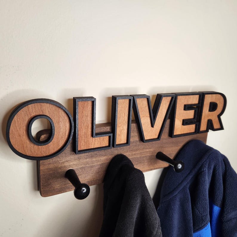 Pu&ograve; includere: Un appendiabiti in legno con il nome "OLIVER" in grandi lettere marroni con contorno nero. L'appendiabiti ha tre ganci in metallo nero ed &egrave; montato su una parete.