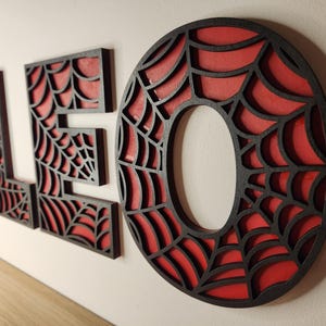 May include: Black wooden letters spelling "LEO" with a red spiderweb design. The letters are cut out and the spiderweb design is visible through the cutouts.