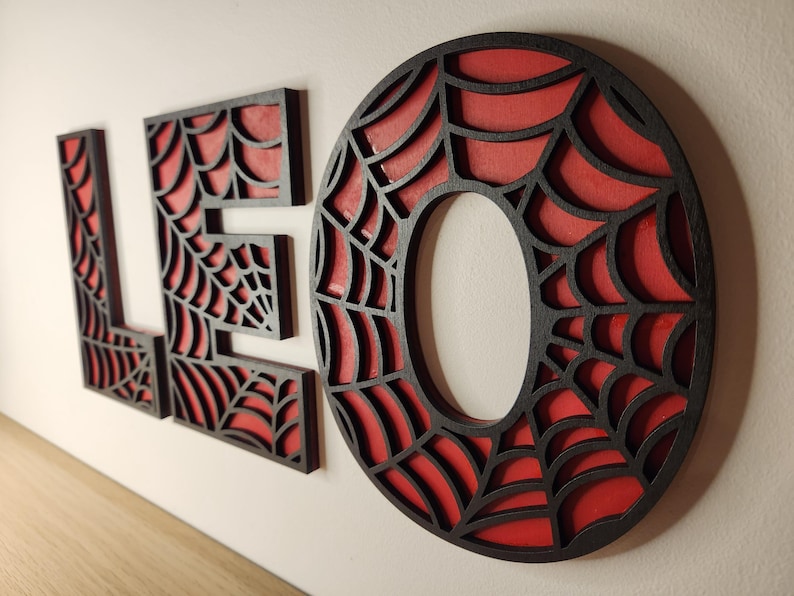 May include: Black wooden letters spelling "LEO" with a red spiderweb design. The letters are cut out and the spiderweb design is visible through the cutouts.