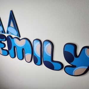 May include: Blue and beige wooden letters spelling "EMILY" with a blue and white geometric pattern. The letters are attached to a white wall.