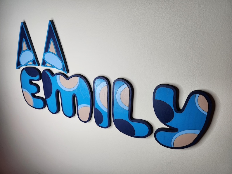 May include: Blue and beige wooden letters spelling "EMILY" with a blue and white geometric pattern. The letters are attached to a white wall.
