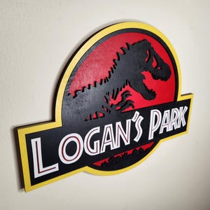 May include: Wooden sign featuring a black dinosaur silhouette against a red background, with the text "LOGAN'S PARK" in white letters. The sign has a yellow border and is mounted on a light-colored wall.