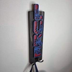 May include: A vertical wooden wall sign featuring the name LUKE in blue, spiderweb-patterned letters layered over a red background. The sign is mounted on a dark gray rectangular wooden board with a black metal hook at the bottom.