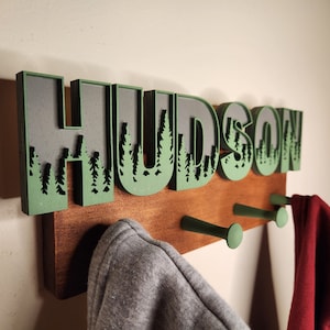 May include: A wooden coat rack with the name "HUDSON" spelled out in green letters with a forest scene design. The rack has three hooks and two hooded sweatshirts hanging on it.