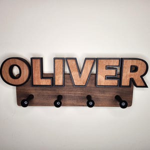 Pu&ograve; includere: Un appendiabiti in legno con il nome "OLIVER" scritto in grandi lettere con contorno nero. L'appendiabiti ha quattro ganci neri.