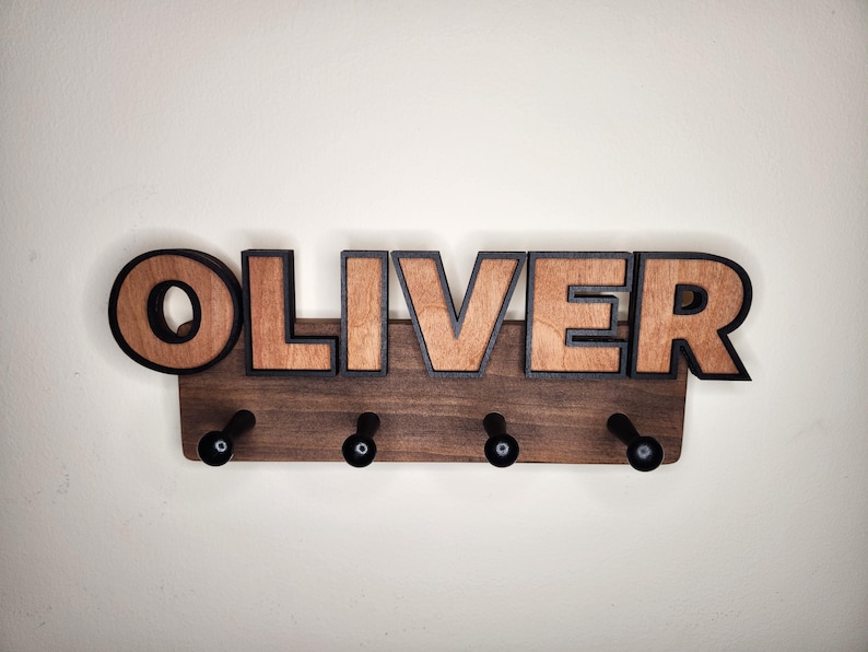 Pu&ograve; includere: Un appendiabiti in legno con il nome "OLIVER" scritto in grandi lettere con contorno nero. L'appendiabiti ha quattro ganci neri.