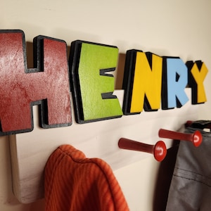 May include: A wooden coat rack with the name "HENRY" spelled out in colorful, 3D letters. The letters are red, green, yellow, blue, and black. There are three red hooks on the rack, with a gray hat and an orange shirt hanging on them.