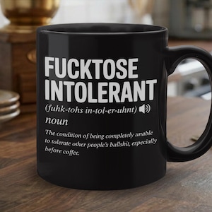 May include: Black ceramic coffee mug with the text "FUCKTOSE INTOLERANT" in white, along with a definition. The mug has a curved handle and is on a wooden surface. The mug is a humorous gift.