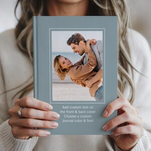 May include: A journal with a pale blue cover and a framed photograph of a couple embracing. The text on the cover reads: "Add custom text on the front & back cover. Choose a custom journal color & font."