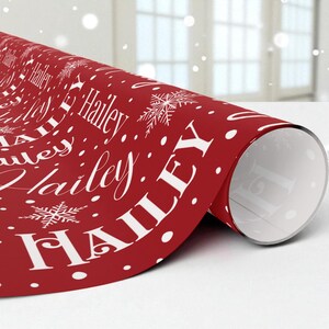 May include: A roll of red wrapping paper with the name "Hailey" in white script and block letters, along with white snowflakes and polka dots. The paper is partially unrolled, revealing the white interior.