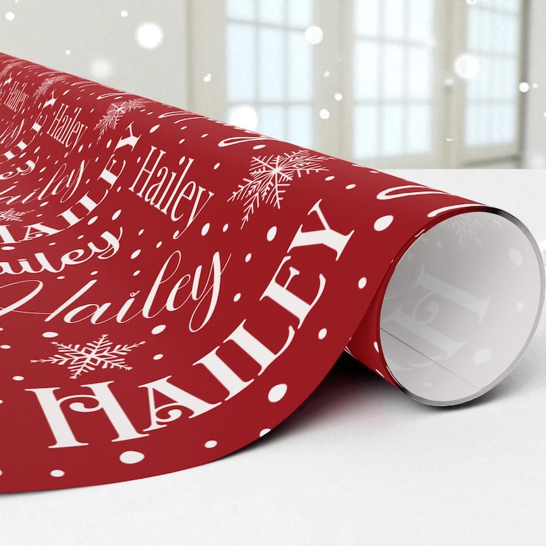 May include: A roll of red wrapping paper with the name "Hailey" in white script and block letters, along with white snowflakes and polka dots. The paper is partially unrolled, revealing the white interior.