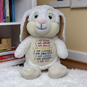 Peut inclure: Une peluche lapin blanche avec de longues oreilles, un nez rose et de grands yeux bleus. Le ventre présente un texte brodé : I AM KIND, I AM SMART, I AM BRAVE, I AM CONFIDENT, I AM CAPABLE, I AM GRATEFUL, I AM LOVED, I AM ENOUGH, I AM IVY.