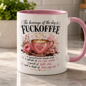 May include: White ceramic coffee mug with a pink interior and handle. The mug features the text "FUCKOFFEE" above a pink coffee cup graphic with floral accents and the text "A special brew made with...kiss my ass."