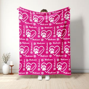 May include: A magenta blanket with a repeating pattern of white hearts containing paw prints and the name "Madison". The blanket also features scattered paw prints and the name "Madison". The blanket is held up by a person.