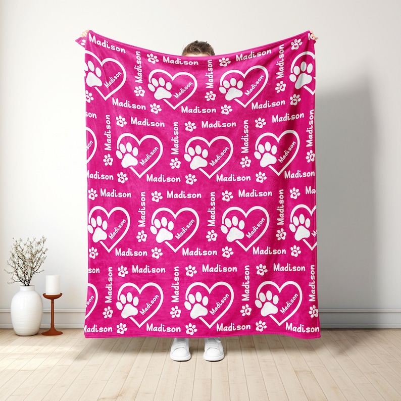 May include: A magenta blanket with a repeating pattern of white hearts containing paw prints and the name "Madison". The blanket also features scattered paw prints and the name "Madison". The blanket is held up by a person.
