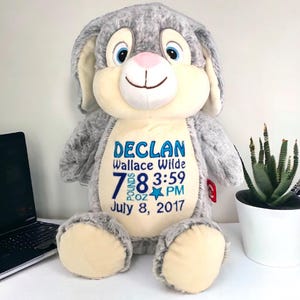 May include: A plush gray bunny rabbit with cream-colored accents. The bunny has blue eyes and a pink nose. Text on the bunny reads "Declan Wallace Wilde" with birth details: 7 lbs 8 oz, 3:59 PM, July 8, 2017.
