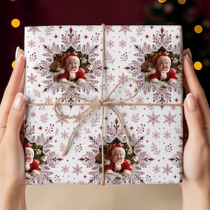 May include: A square gift box wrapped in white paper featuring a repeating pattern of red snowflakes, sprigs, and stars. The paper includes three circular photo inserts of a baby wearing a red Santa suit and hat, tied with thin brown twine.
