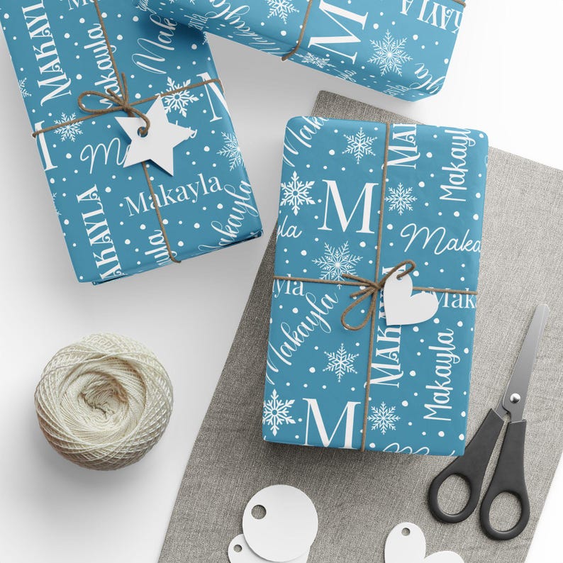 May include: Gift-wrapped presents with blue wrapping paper featuring white snowflakes, polka dots, and the name "Makayla." Each package is tied with twine and a white tag. A ball of twine, scissors, and blank tags are also visible.