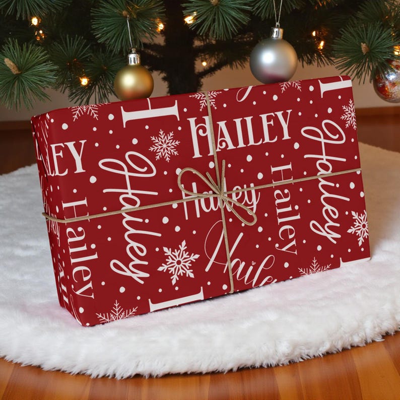 May include: A wrapped gift with a red background and white text that reads "Hailey" in a repeating pattern. The gift is tied with twine and decorated with snowflakes and white dots. The gift is placed on a white fluffy surface.