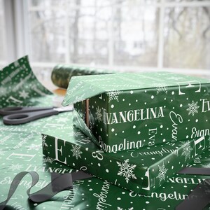 May include: Two wrapped gifts with green paper featuring the name "Evangelina" in white, along with snowflakes and polka dots. A pair of scissors and a roll of matching wrapping paper are visible.