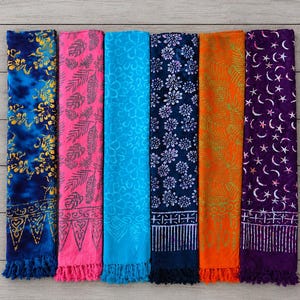 May include: Six rectangular sarongs with fringe edges, displayed in a row. The fabrics feature various patterns including gold floral prints on blue, pink leaf motifs, light blue flowers, dark blue daisies, orange foliage, and purple moon and stars.