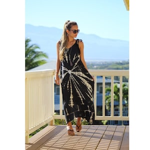 May include: A black and white tie-dye maxi dress with a halter neckline and tie straps. The dress features a circular design at the waist and is worn with sandals. The background includes a wooden deck, a white railing, and a scenic view.