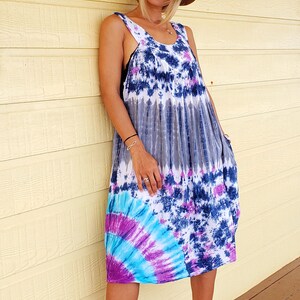 May include: A woman wearing a tie-dye maxi dress with a brown hat and brown wedge sandals. The dress has shades of blue, purple, and white.
