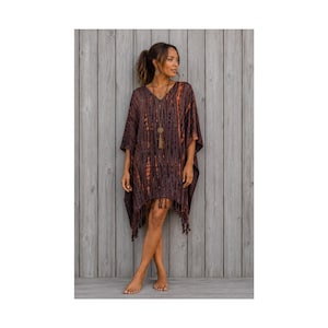 May include: A dark brown and orange tie-dye poncho featuring a V-neckline, short wide sleeves, and a fringed hemline. The garment has a vertical striped pattern created by the tie-dye process and hangs loosely over the body to the mid-thigh.