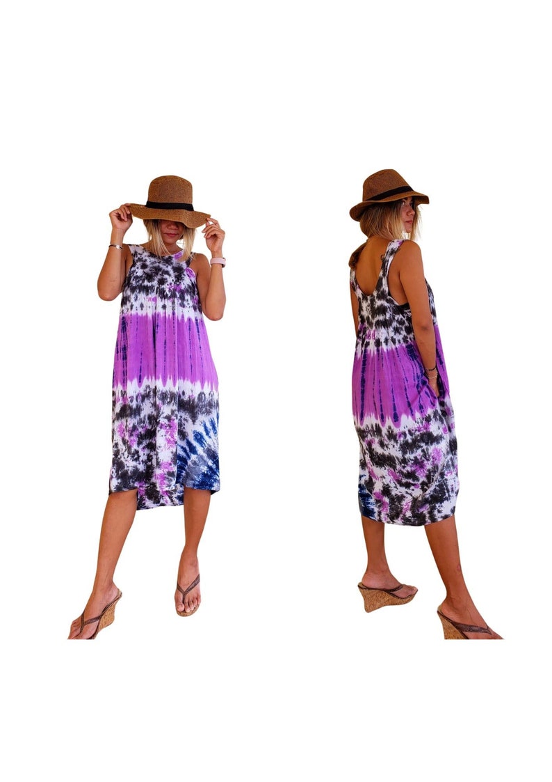 K&ouml;nnte beinhalten: Ein &auml;rmelloses Kleid in Lila, Schwarz und Wei&szlig; mit Batik-Muster und einem runden Ausschnitt. Das Kleid ist knielang und hat eine lockere Passform. Das Model tr&auml;gt einen braunen Hut und braune Sandalen.