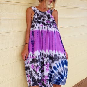 K&ouml;nnte beinhalten: Eine Frau tr&auml;gt ein lila, schwarz und wei&szlig;es Batik-Kleid mit einem braunen Fedora-Hut und braunen Sandalen.