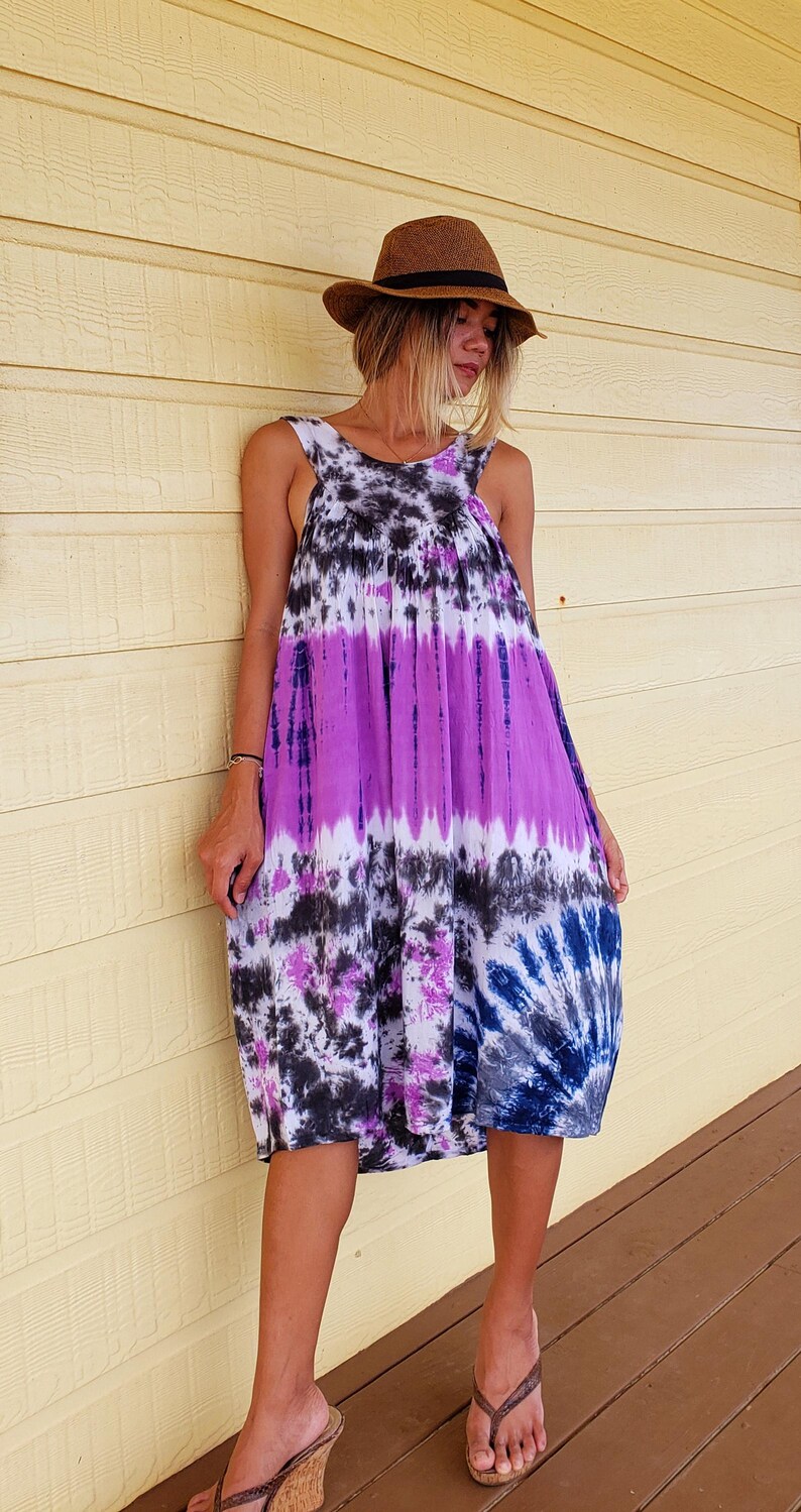 K&ouml;nnte beinhalten: Eine Frau tr&auml;gt ein lila, schwarz und wei&szlig;es Batik-Kleid mit einem braunen Fedora-Hut und braunen Sandalen.
