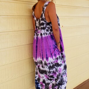 K&ouml;nnte beinhalten: Eine Frau tr&auml;gt ein lila, schwarz und wei&szlig;es tie-dye-Kleid ohne &Auml;rmel mit einem braunen Hut und braunen Keilsandalen.