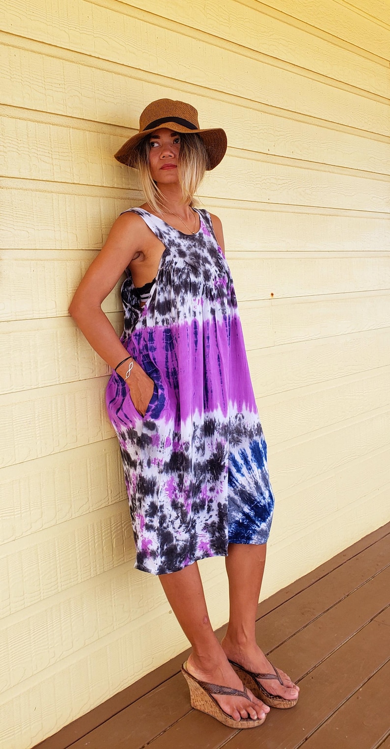 K&ouml;nnte beinhalten: Eine Frau tr&auml;gt ein lila, schwarz und wei&szlig;es Batik-Kleid mit einem braunen Hut mit breiter Krempe und braunen Keilsandalen.