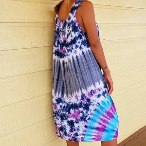 May include: A woman wearing a tie-dye maxi dress with a brown hat and brown wedge sandals. The dress has shades of blue, purple, and white.