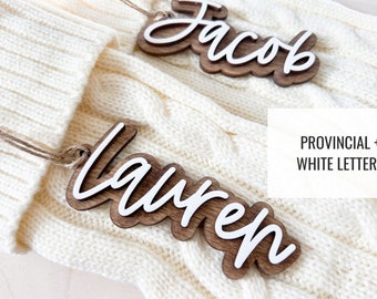 Puede incluir: Dos adornos de madera con los nombres "Jacob" y "Lauren" escritos en letras blancas. Los adornos están sobre una superficie de punto blanco. El texto "Provincial + White Letters" es visible en el fondo.