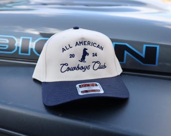 May include: A beige and navy blue baseball cap with the text "All American 20 24 Cowboys Club" embroidered on the front. A silhouette of a horse is also embroidered on the front.