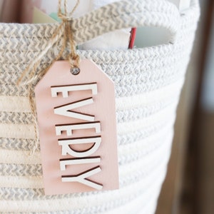 May include: A pink wooden gift tag with the word "EVERYDAY" written in white letters. The tag is tied to a gray and white striped rope basket.