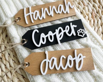 Op de afbeelding: Drie houten cadeau-etiketten met namen in witte schrijfletters. De etiketten zijn vastgebonden met touw en hebben een natuurlijke houten afwerking. De namen op de etiketten zijn "Hannah", "Cooper" en "Jacob".