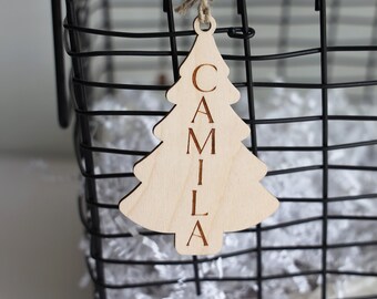 Könnte beinhalten: Ein hölzerner Weihnachtsbaum-Anhänger mit dem eingravierten Namen "Camila". Der Anhänger hängt an einem Drahtkorb, der mit weißem zerknülltem Papier gefüllt ist.