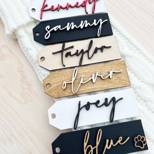 Puede incluir: Seis etiquetas de regalo de madera con nombres escritos en pintura negra y roja. Los nombres son "Kennedy", "Sammy", "Taylor", "Oliver", "Joey" y "Blue". La última etiqueta tiene una pequeña huella de pata.